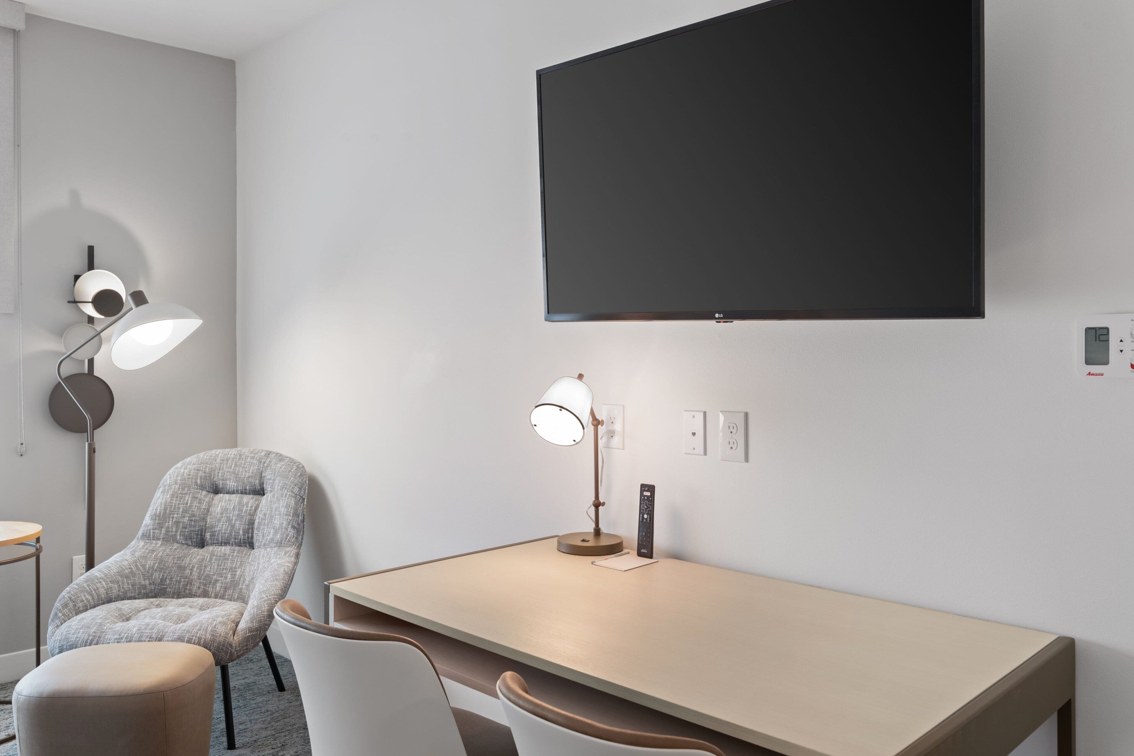 A modern hotel room with a flat-screen TV mounted on the wall, a desk with a lamp, and a comfortable armchair.