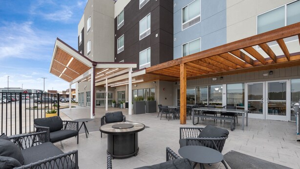 An outdoor seating area with wicker chairs and tables, a wooden pergola, and a building with large windows and a flat roof.