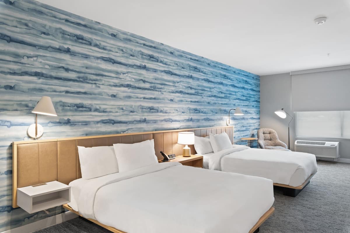 A hotel room with two beds, a large blue patterned wall, a window with blinds, and a small table with a lamp.