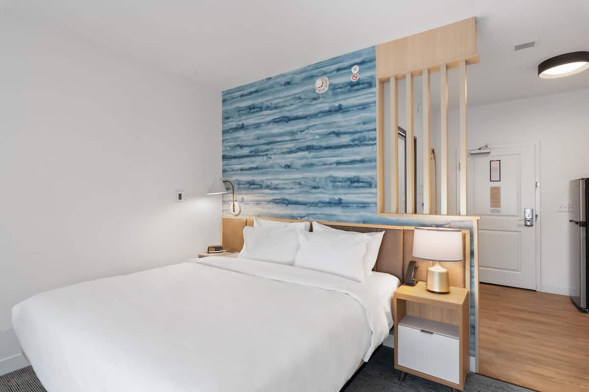A modern hotel room with a large bed, a nightstand with a lamp, a small bedside table, and a wall-mounted artwork.