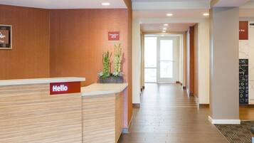 Zona de recepción de hotel con un mostrador de recepción que muestra la leyenda “Hello”, un cartel con los rótulos “Stay”, “Work” y “Play”, un mostrador, una planta y un mapa en la pared.