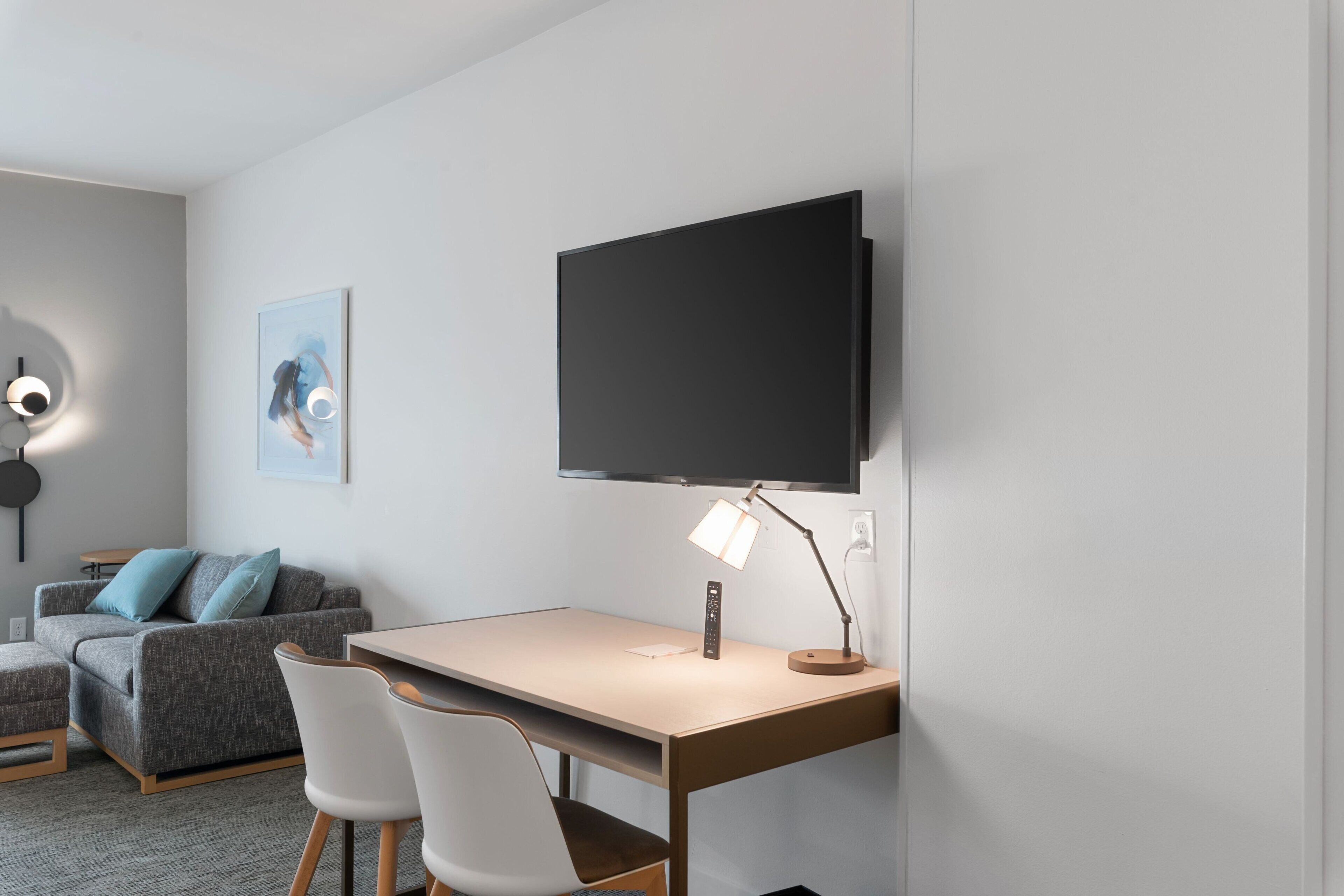 A modern room with a desk, a flat-screen TV mounted on the wall, a desk lamp, a couch, and a painting.