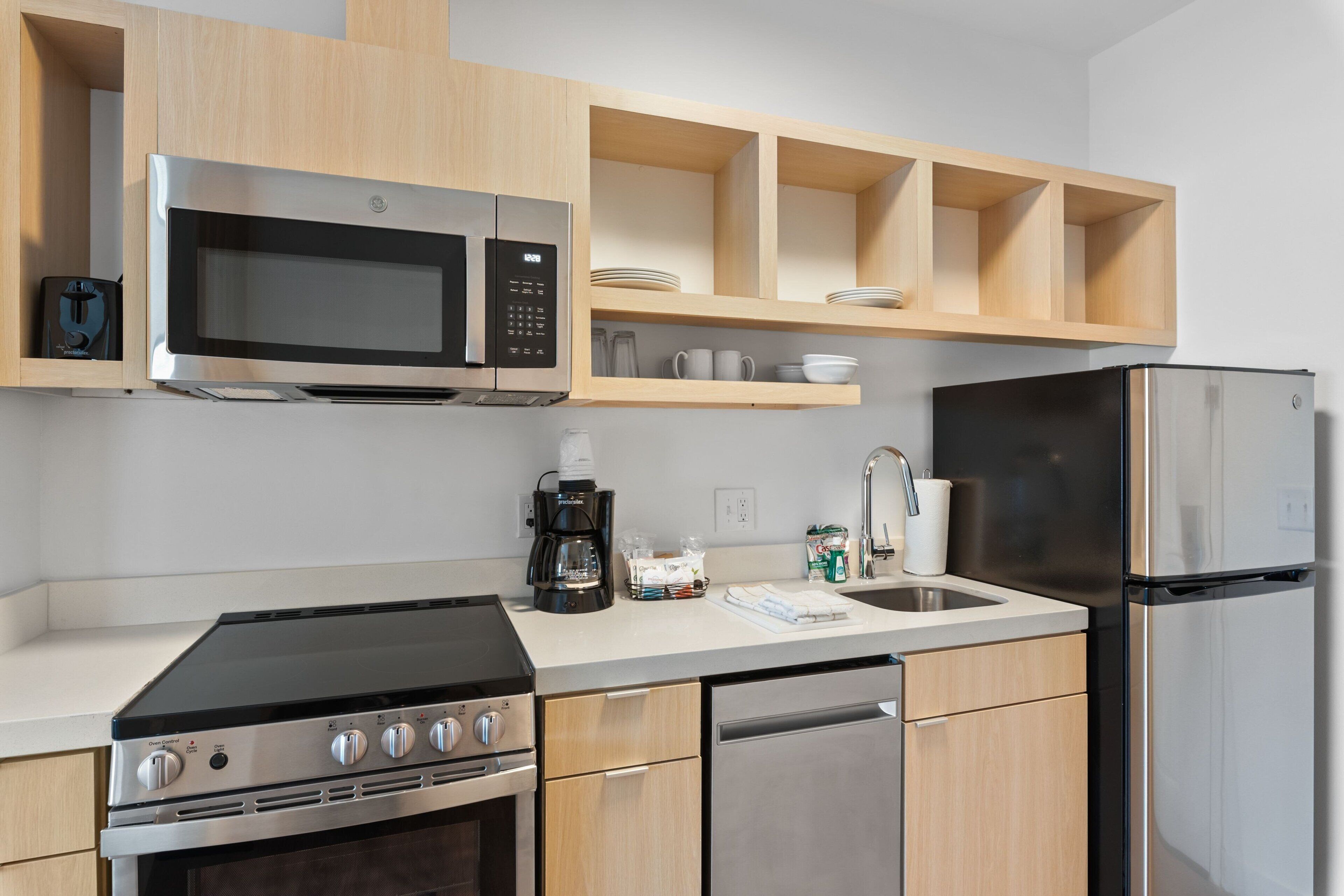 A modern kitchen with a built-in microwave, stove, and dishwasher. There are wooden cabinets and a countertop with a coffee maker and cups.