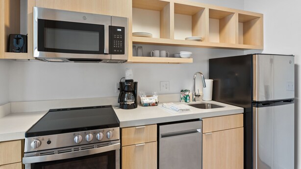 Una cocina moderna con microondas, estufa y lavavajillas integrados. Cuenta con armarios de madera y una encimera con cafetera y tazas.