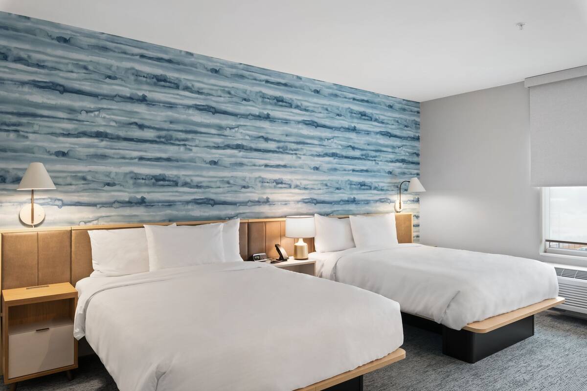 A hotel room with two beds, a large blue abstract wall, bedside lamps, and a window with blinds.