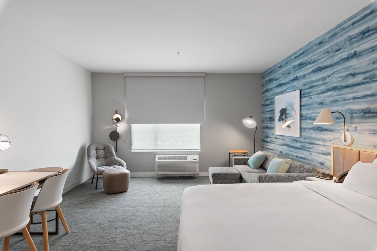 A modern hotel room with a bed, a desk, a chair, a sofa, a small table, and a wall with a blue abstract design.