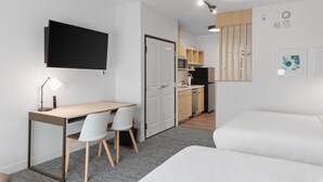 A modern, compact living space with a kitchenette, a flat-screen TV mounted on the wall, a desk with a chair, and a bed.