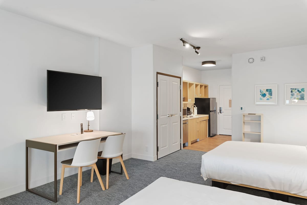 A modern hotel room with a desk, two chairs, a bed, a TV, and a kitchenette in the background.