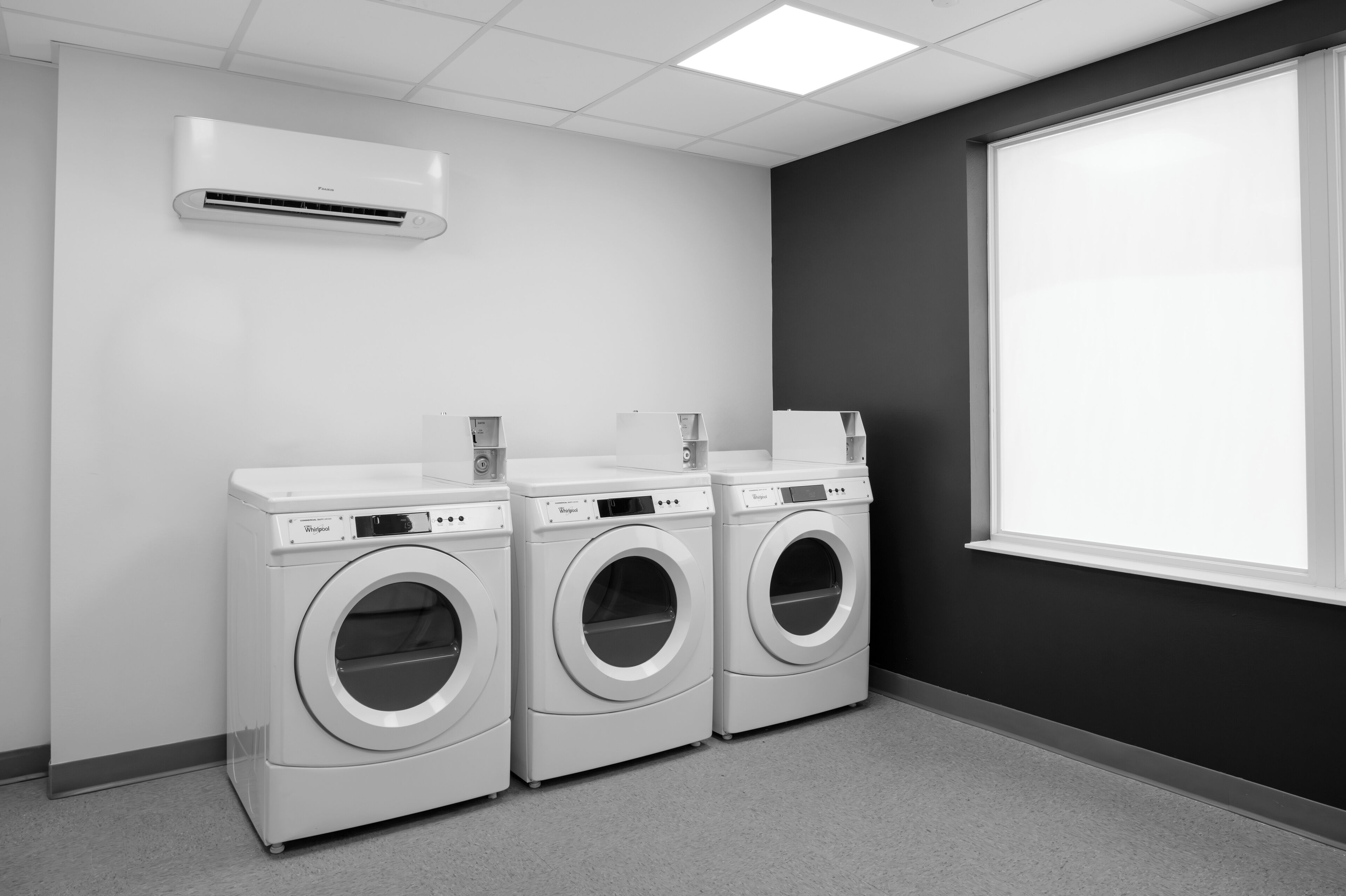 A laundry room with three white front-loading washers and dryers, a white air conditioning unit, and a large window.