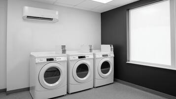 A laundry room with three white front-loading washers and dryers, a white air conditioning unit, and a large window.