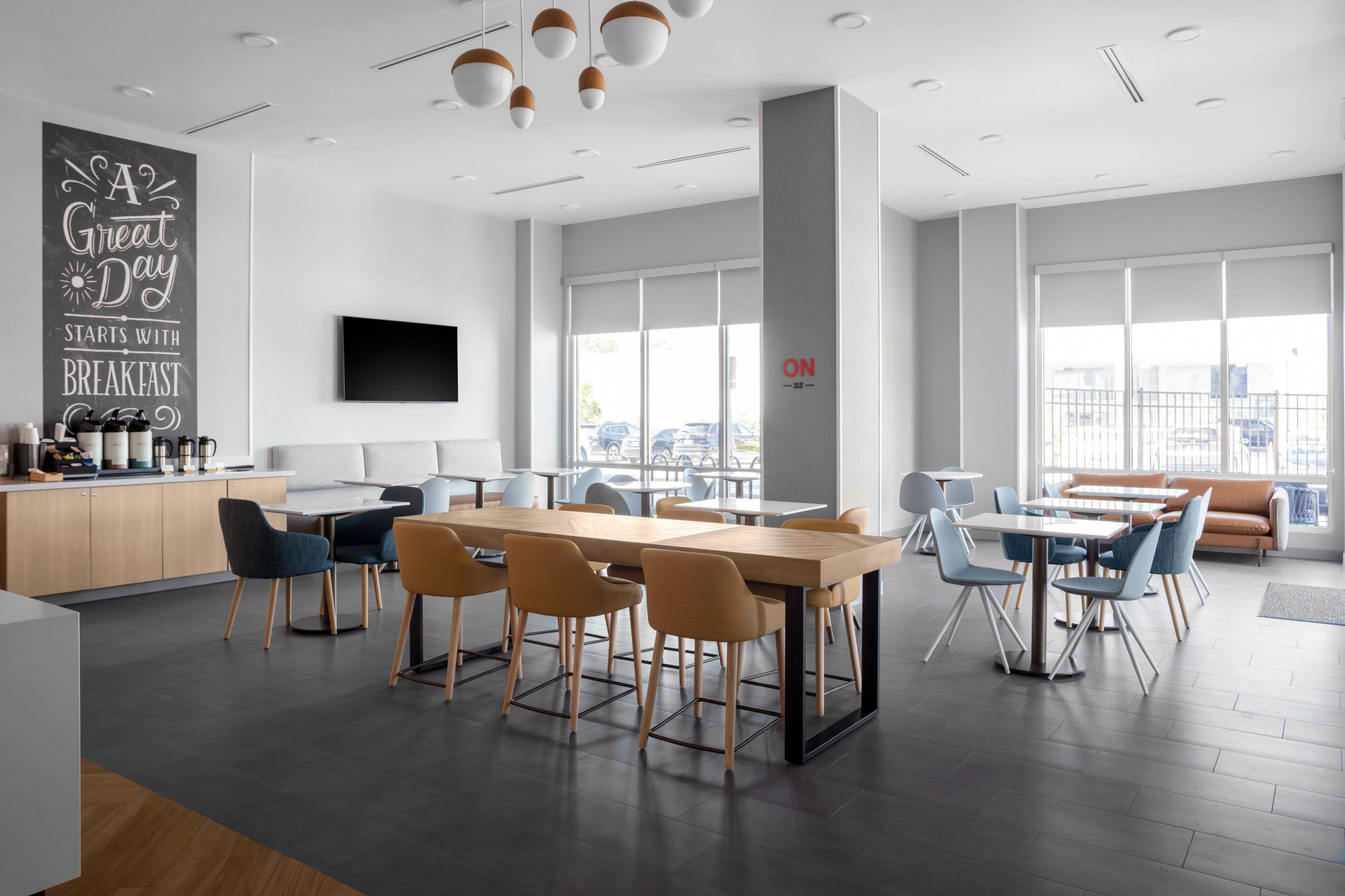 A modern dining area with a long wooden table, blue and orange chairs, and a kitchen area with a coffee station.