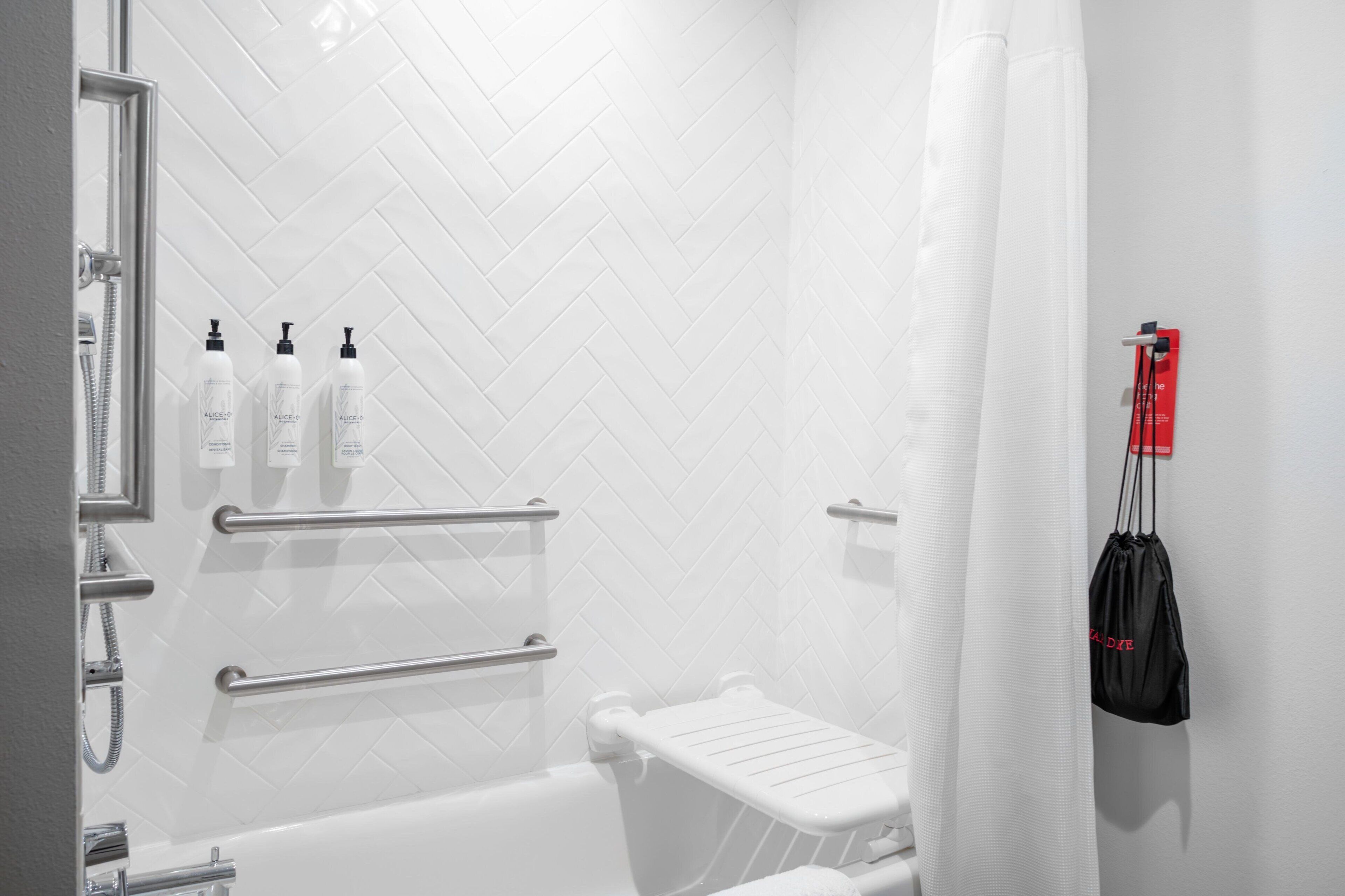 A modern bathroom with white herringbone tiles, a white bathtub, and a shower area with a glass door. There are three bottles of shampoo and a black bag hanging on the wall.