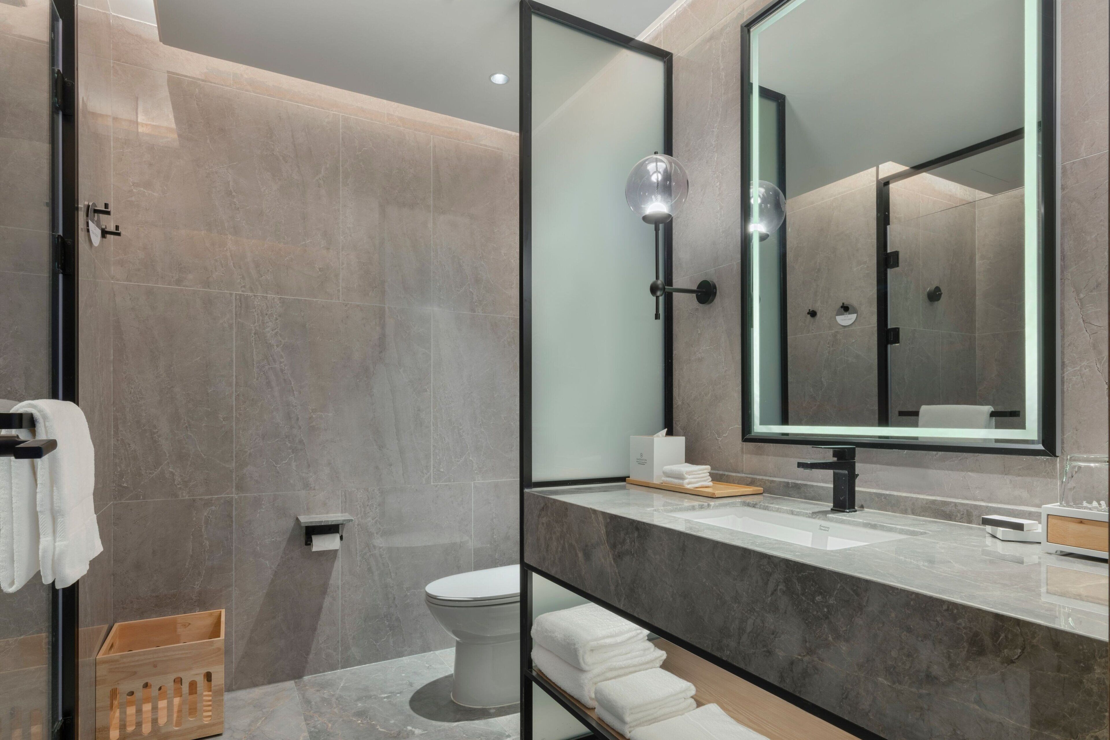 A modern bathroom with a glass shower enclosure, a large mirror, a marble countertop, and a toilet.