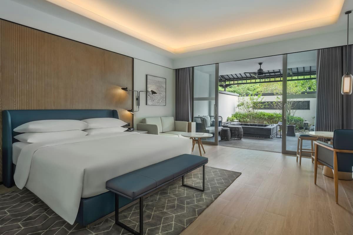 A modern hotel room with a large bed, a bench, a small table, and a seating area with a view of the outdoors.