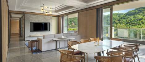 A spacious living area with a large TV, comfortable seating, a dining table, and a balcony with a scenic view.
