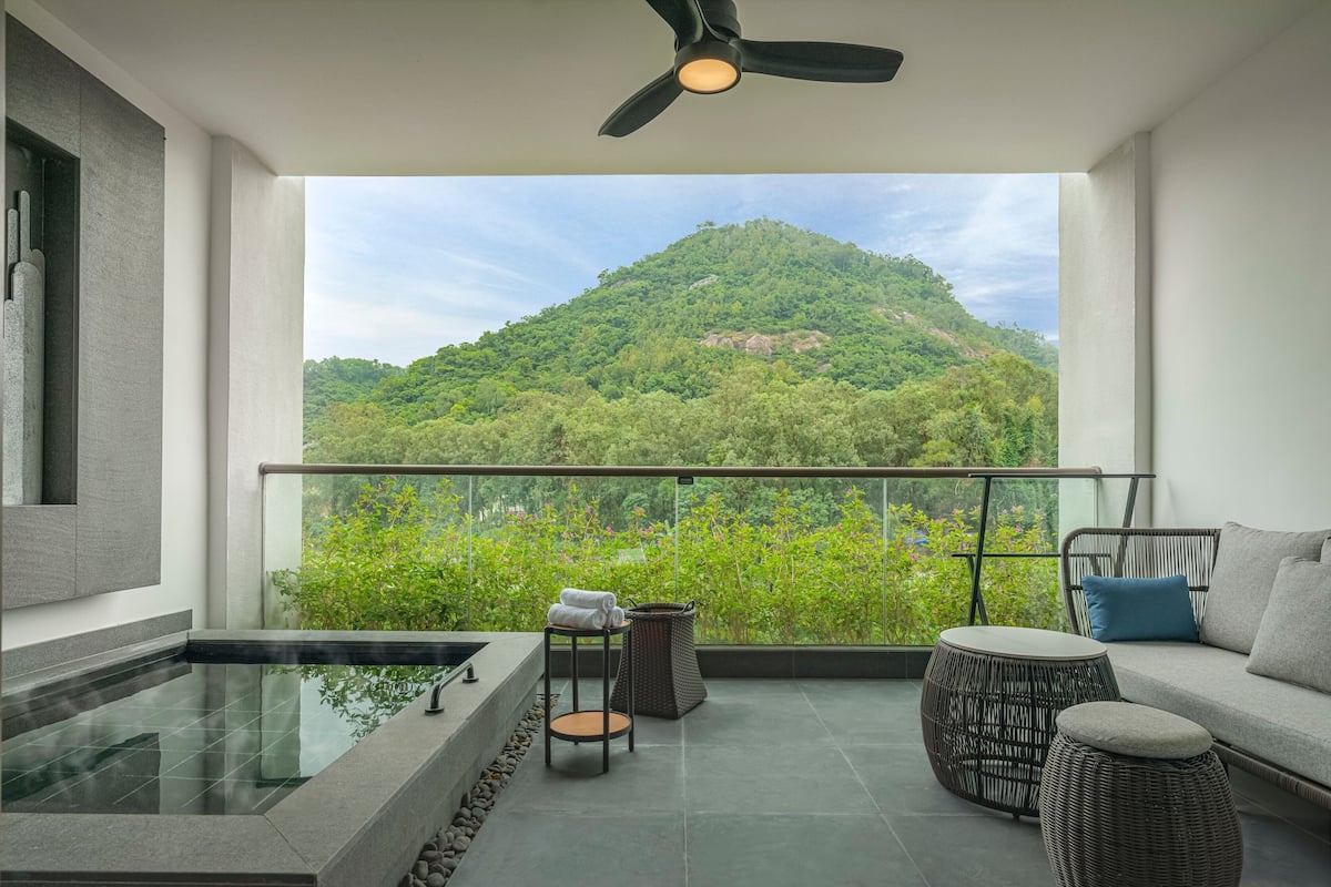 A balcony with a view of a green hill, a seating area with a sofa and a table, and a small hot tub.