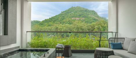 Un balcón con vistas a una colina verde, una zona de estar con sofá y mesa, y un jacuzzi pequeño.