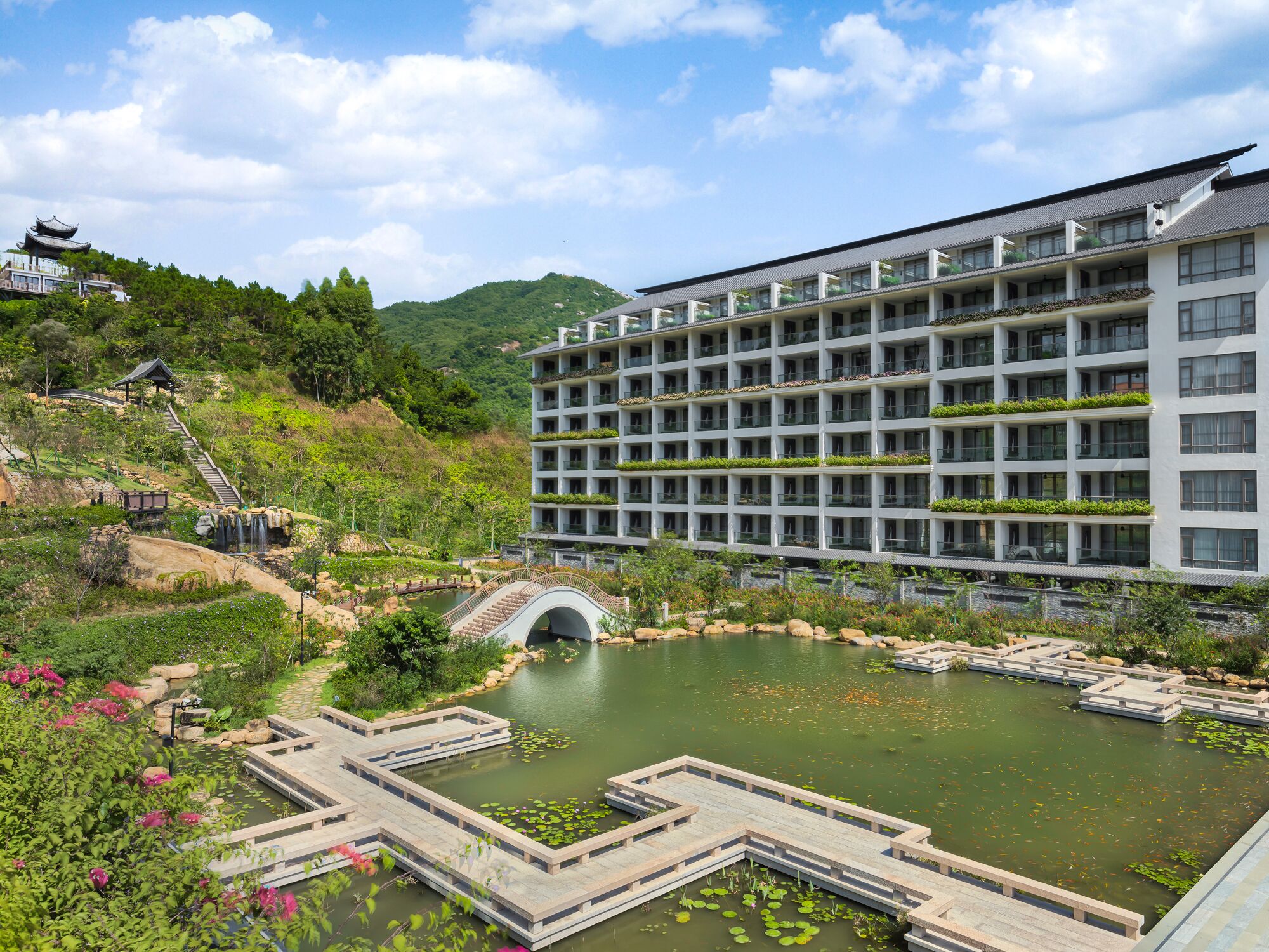 A hotel with a modern design, featuring multiple balconies with plants, a large central courtyard with a pond, and a traditional-style building in the background.