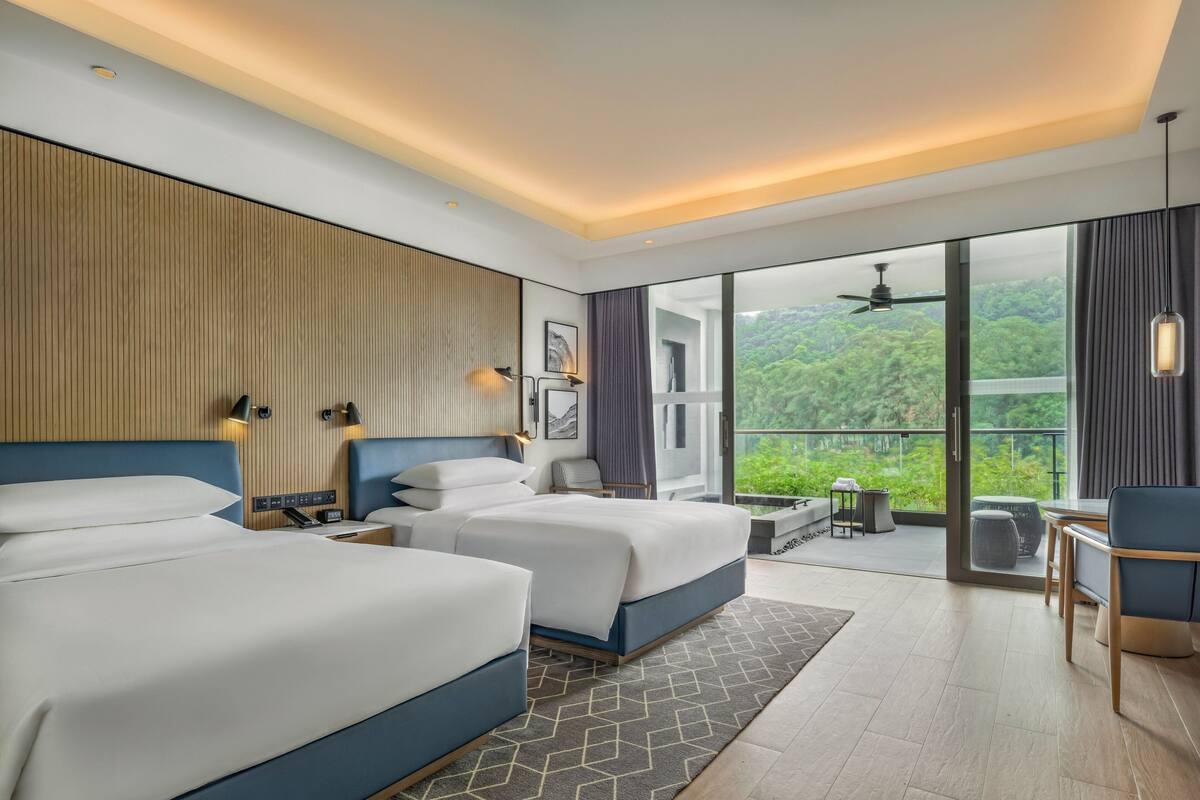 A modern hotel room with two beds, a balcony with a view of greenery, and a seating area with a table and chairs.