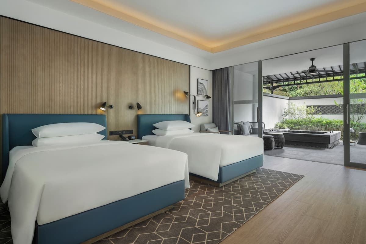 A modern hotel room with two beds, a wooden accent wall, and a view of a garden through large glass doors.