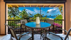 Une table ronde, entourée de chaises, et un vase sont disposés sur un balcon qui offre une vue imprenable sur un paysage tropical, avec des palmiers et l’océan à l’horizon.