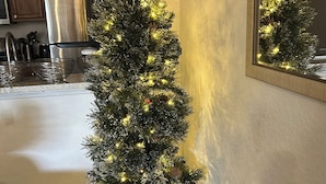 Un arbre de Nadal en una test de color blanc amb llums, col·locat en una habitació amb terra de fusta, un llit i una cuina amb armaris i electrodomèstics.