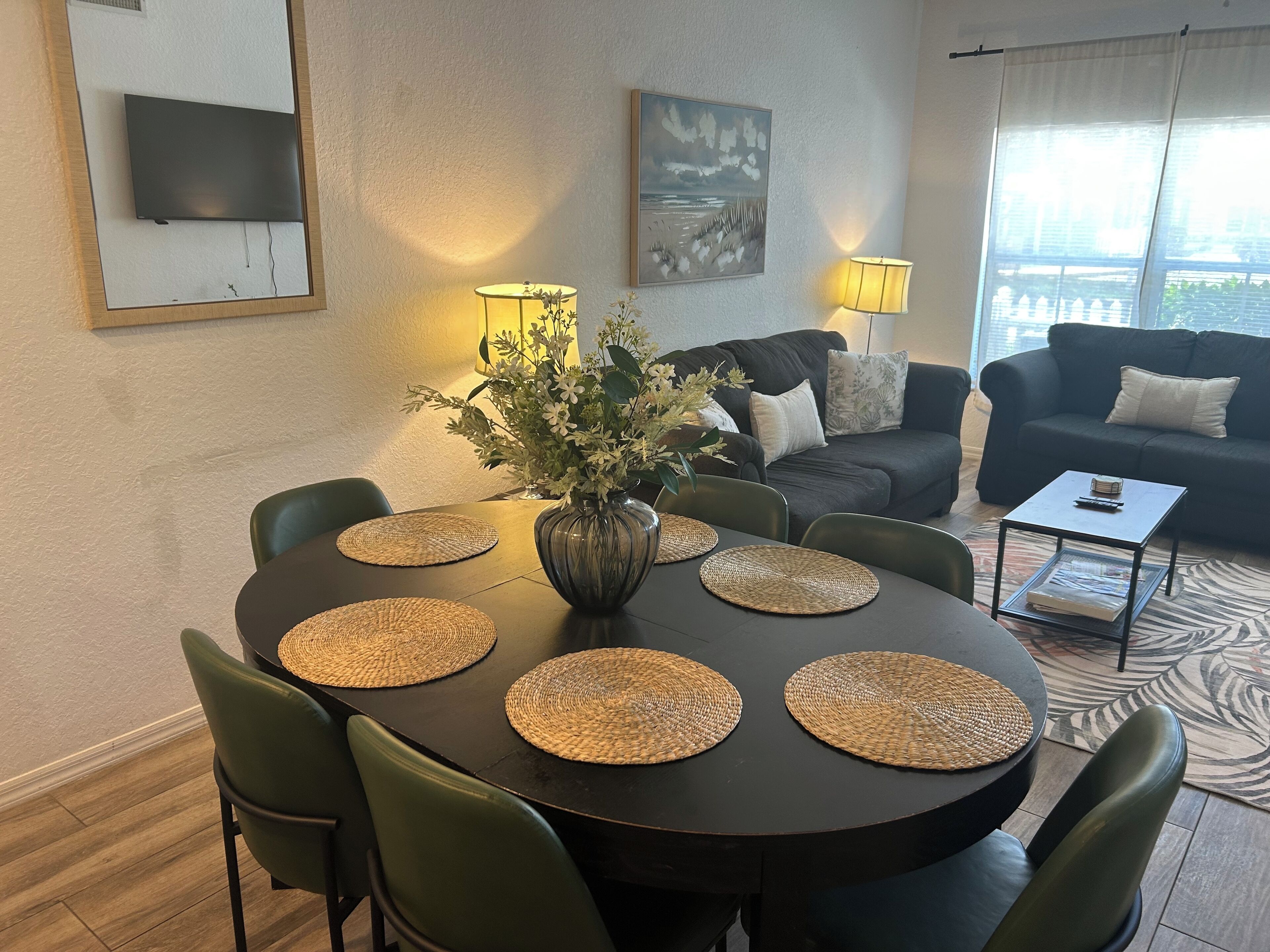 Ein Essbereich mit einem runden Tisch, grünen Stühlen, gewobenen Tischsets, einer Vase mit Blumen und einer Couch im Hintergrund.