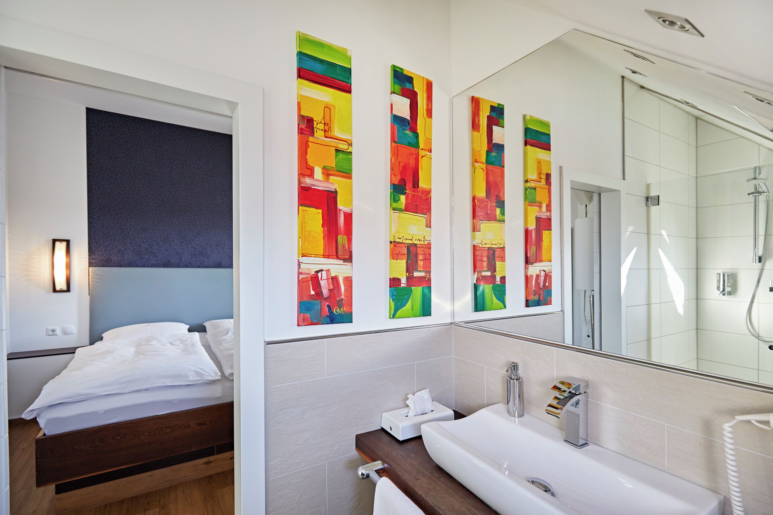 Habitación de hotel con cama, baño con lavamanos y espejo, y arte colorido en la pared.