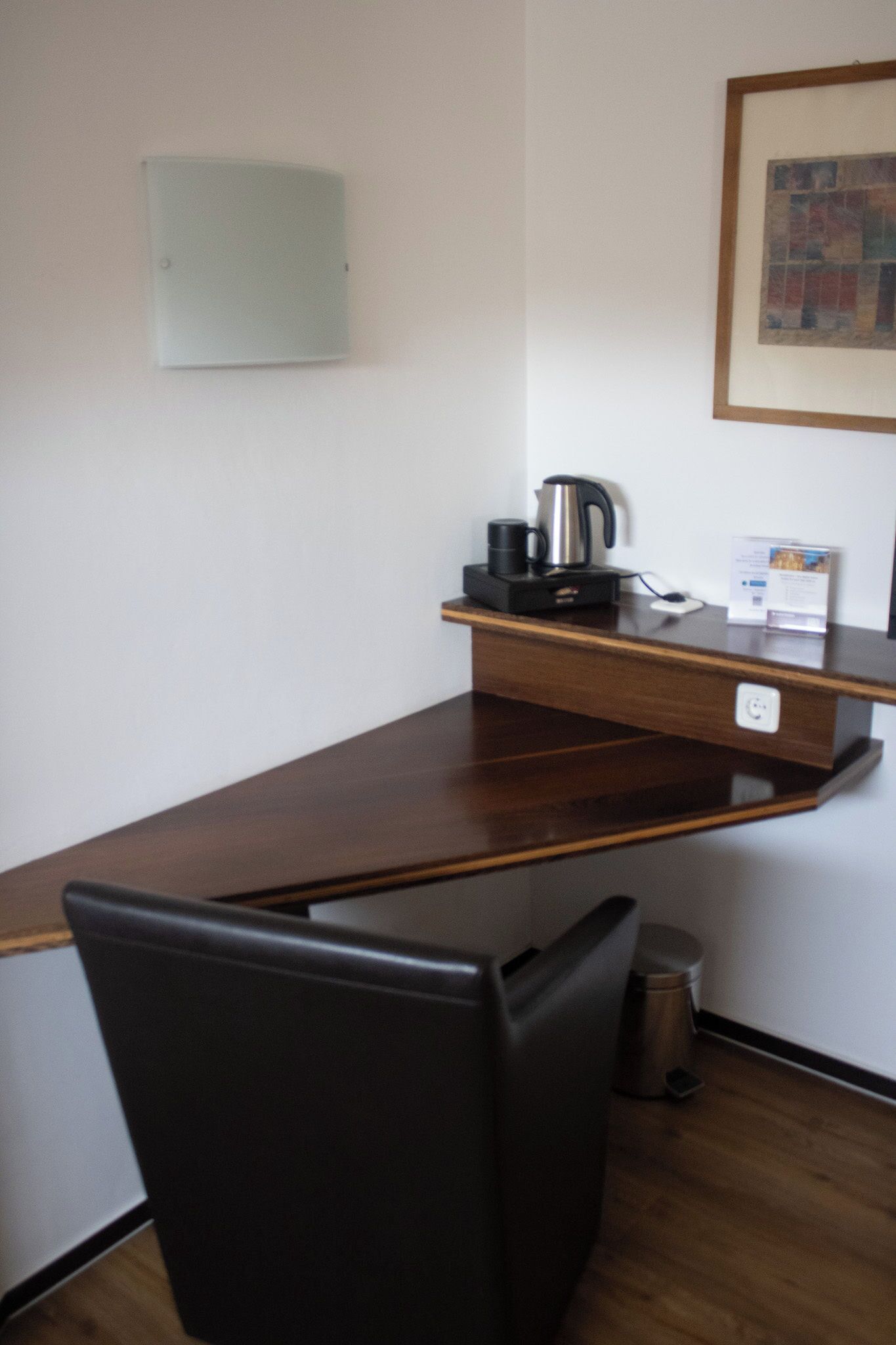 A small office space with a wooden desk, a black chair, a coffee maker, and a framed picture on the wall.