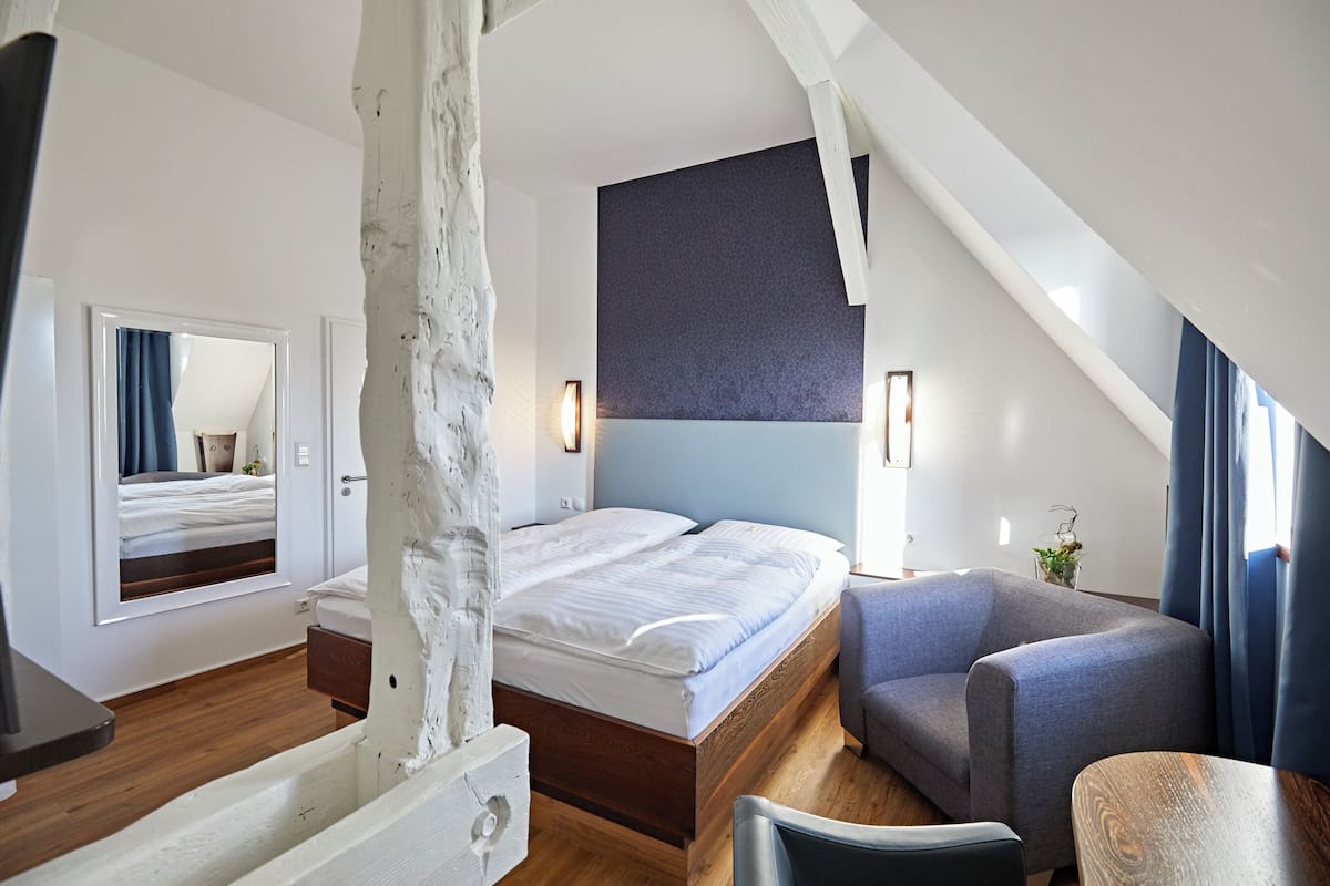 Ett modernt hotellrum med en säng i trä, en grå fåtölj och ett litet bord.