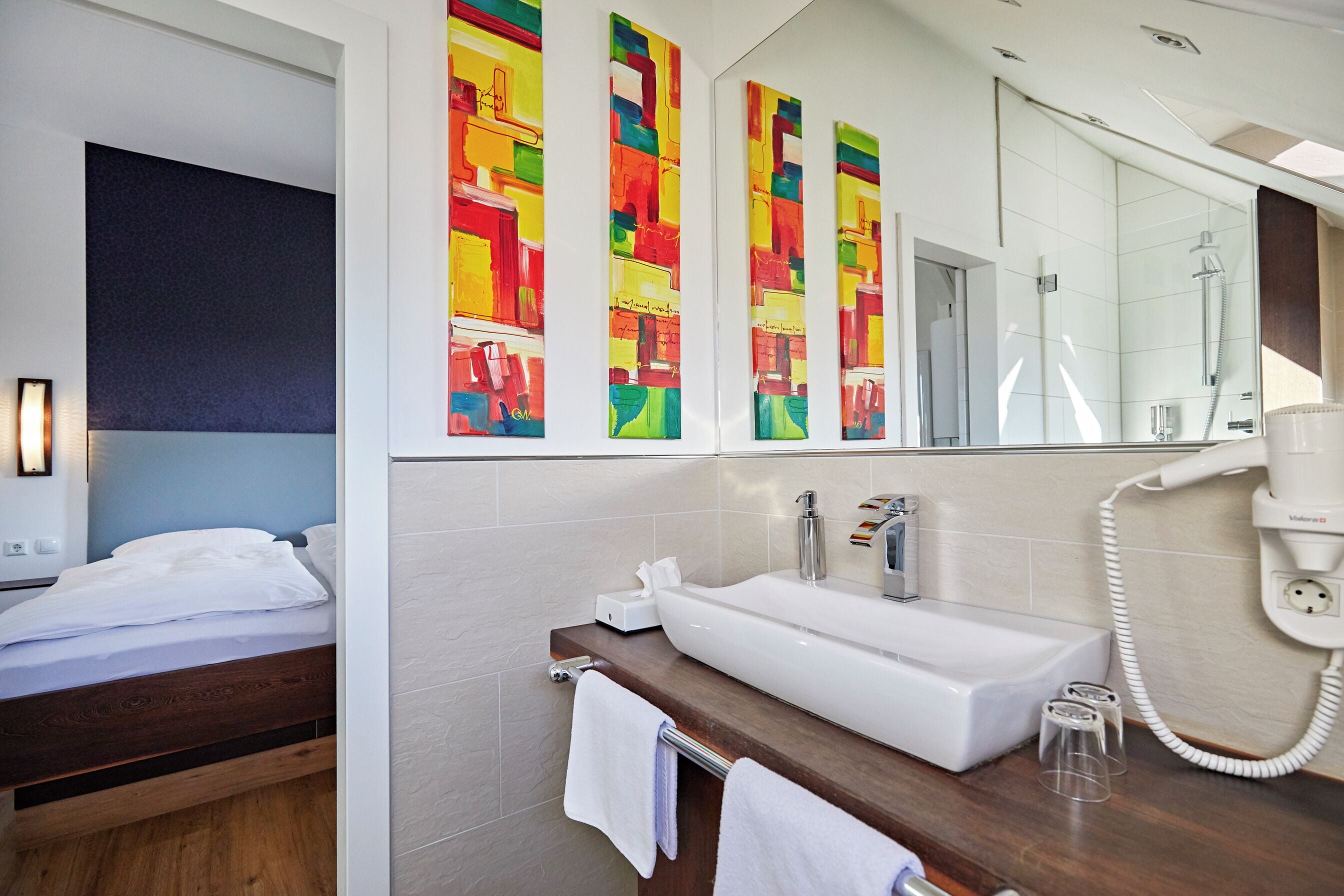 A modern bathroom with a large mirror, a white sink, and a shower. There are colorful artwork pieces on the wall and a wooden bed in the adjacent room.