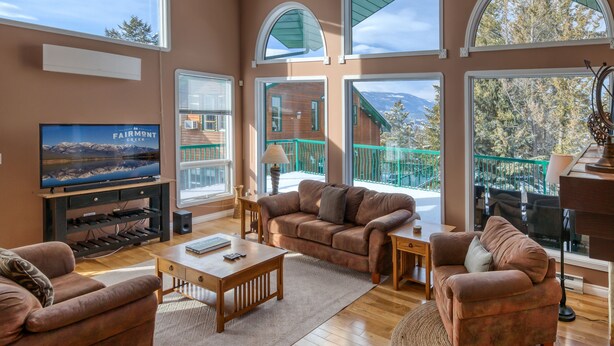 A spacious living room with a large TV, comfortable seating, and large windows offering views of a mountain and a building. - Marble Canyon 3 Bdrm|Sleeps 8 (12 inc kids): Unit 102P1 (Fairmont Hot Springs)