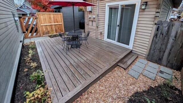 A wooden deck with a table and chairs, a red umbrella, and a sliding glass door. - Bluebird Day - River Otter, Charming 1BR duplex in Old Downtown Bend (Bend)