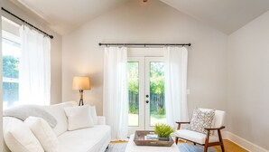 Een woonkamer met een witte bank, een houten salontafel en een gestreept fauteuil. Er staat een lamp op een bijzettafeltje en er is uitzicht door een glazen deur.