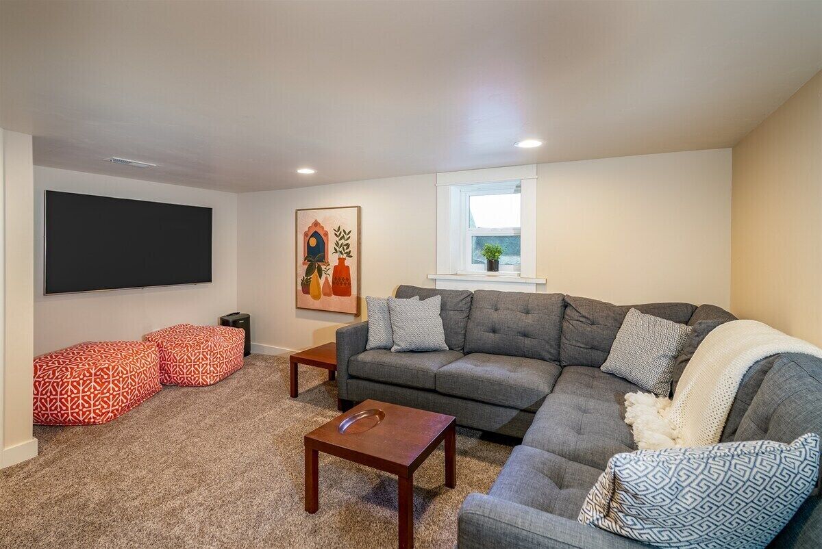 Ein Wohnzimmer mit einem grauen Ecksofa, einem Couchtisch und zwei Hocker mit geometrischem Muster.