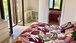 Un dormitorio con cama, una puerta corrediza de vidrio que da a un balcón, un baño pequeño y una ventana con vistas a la vegetación.