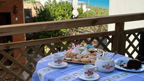 Una mesa dispuesta con pasteles, tazas de té y un pastel, en un balcón con vista al mar.
