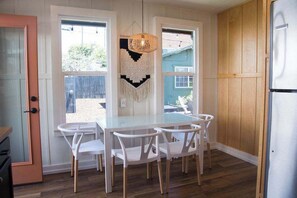 A kitchen with a glass table, white chairs, a hanging light fixture, and a wooden cabinet. - 1915 Boho California Beach Kit House (Oceanside)