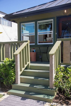 A house with a green exterior, a set of stairs leading to a porch, a small table with a chair, and a window with a laptop on it. - 1915 Boho California Beach Kit House (Oceanside)