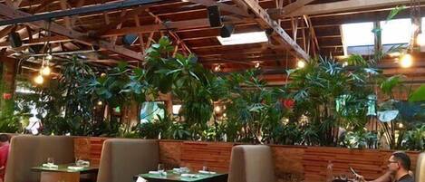 An indoor restaurant with wooden beams, tables, chairs, and potted plants.