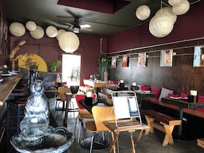 A restaurant interior with wooden tables and chairs, a central seating area with red cushions, and hanging paper lanterns. - 1915 Boho California Beach Kit House (Oceanside)