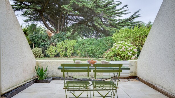 A patio with a table and chairs, surrounded by greenery and a large tree. - Le Loriot - One Bedroom Apartment, Sleeps 4 (Saint-Nic)