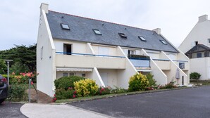 A two-story residential building with a sloped roof, a balcony, and a small garden with flowers. - Le Loriot - One Bedroom Apartment, Sleeps 4 (Saint-Nic)