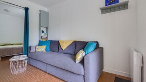 A room with a grey sofa, a small white side table, a bed with green bedding, a blue curtain, a white radiator, and a shelf with a picture. - Le Loriot - One Bedroom Apartment, Sleeps 4 (Saint-Nic)