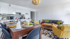 A modern kitchen with white cabinets, a dining area with a wooden table and chairs, and a living room with a sofa and patterned cushions. - L'Airelle - Two Bedroom Apartment, Sleeps 4 (Saint-Malo)