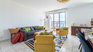 A modern living room with a sofa, two armchairs, a coffee table, and a dining table. There is a large window with a view of the outside, a ceiling light, and a decorative wall hanging. - L'Airelle - Two Bedroom Apartment, Sleeps 4 (Saint-Malo)