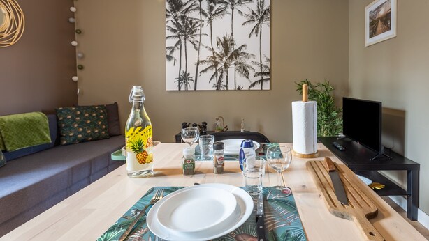 A dining area with a table set for two, featuring a bottle of drink, plates, cutlery, and a painting of palm trees on the wall. - Rochebonne - One Bedroom Apartment, Sleeps 4 (Saint-Coulomb)