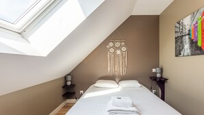 A modern bedroom with a skylight, a bed with white linens, a wall-mounted macrame decoration, and a colorful artwork. - Chevrets - One Bedroom Apartment, Sleeps 2 (Saint-Coulomb)