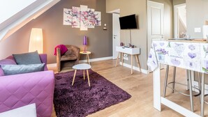 A cozy living room with a purple sofa, a small table, a purple rug, a TV, and a wall with artwork. - Chevrets - One Bedroom Apartment, Sleeps 2 (Saint-Coulomb)