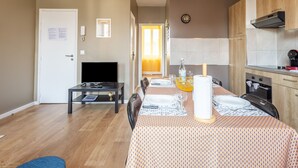 A modern living room with a TV, dining table set for four, and a kitchen with wooden cabinets and a stove. - Minihic - appartement avec jardin (Saint-Coulomb)